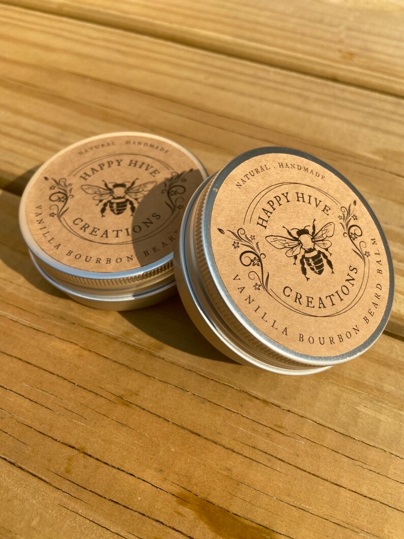 May include: Two small metal tins with brown paper labels. The labels feature a bee illustration and the text "Happy Hive Creations" with the words "Vanilla Bourbon Beard Balm" printed on the bottom of the label.