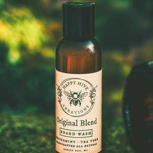 May include: A brown glass bottle with a black cap and a white label. The label says "Happy Hive Creations Original Blend Beard Wash Peppermint Tea Tree Handcrafted All Natural Green Bay, WI".