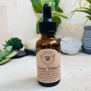 May include: A brown glass bottle with a black dropper top and a brown paper label. The label says "Happy Hive Creations" and "Cherry Tobacco Beard Oil" with a bee illustration. The label also says "Handcrafted All Natural Green Bay, WI".
