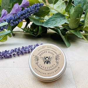 K&ouml;nnte beinhalten: Runde, silberne Dose mit der Aufschrift "Happy Hive Creations Headache Help Balm" und einem Bienen-Logo. Die Dose liegt auf einer hellen Oberfl&auml;che, umgeben von Lavendelzweigen und gr&uuml;nem Laub.