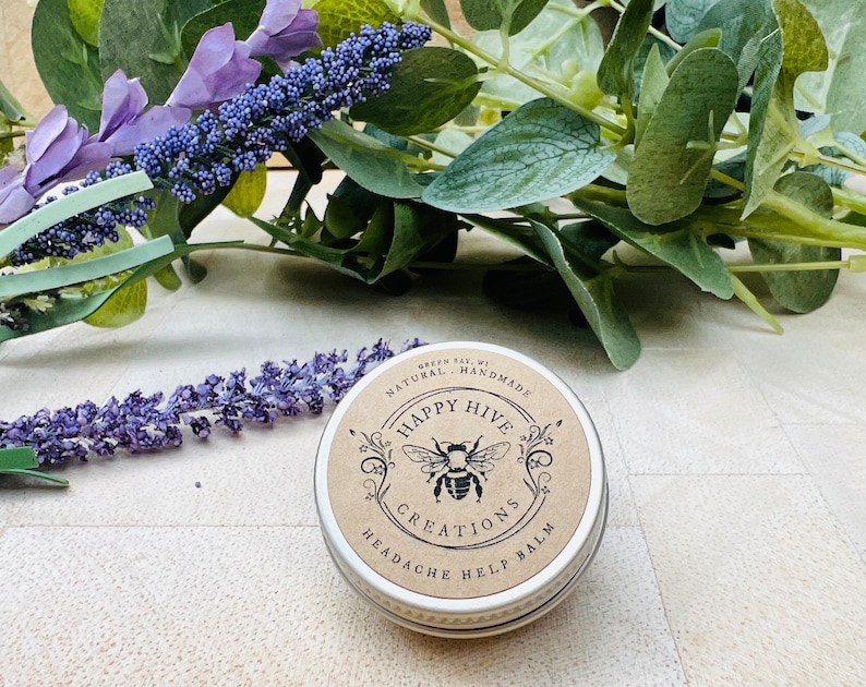K&ouml;nnte beinhalten: Runde, silberne Dose mit der Aufschrift "Happy Hive Creations Headache Help Balm" und einem Bienen-Logo. Die Dose liegt auf einer hellen Oberfl&auml;che, umgeben von Lavendelzweigen und gr&uuml;nem Laub.