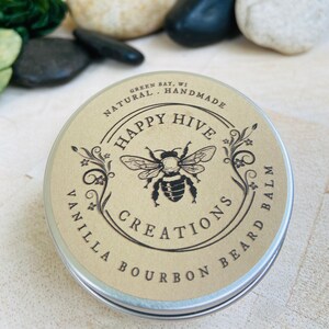 May include: A round metal tin with a brown label. The label has a black and white illustration of a bee with the text "Happy Hive Creations" and "Vanilla Bourbon Beard Balm". The text "Green Bay, WI Handmade Natural" is also on the label.