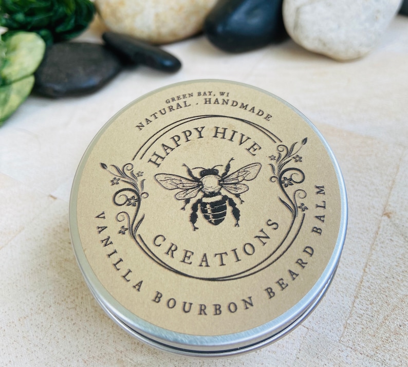May include: A round metal tin with a brown label. The label has a black and white illustration of a bee with the text "Happy Hive Creations" and "Vanilla Bourbon Beard Balm". The text "Green Bay, WI Handmade Natural" is also on the label.