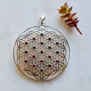 Puede incluir: Un colgante de plata con un diseño de flor de la vida con piedras de granate rojo.