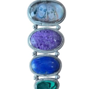 May include: A silver pendant featuring four gemstones. The top gemstone is a clear oval with a painted image of two people. The second gemstone is a purple oval, the third is a blue oval, and the bottom gemstone is a green teardrop shape.