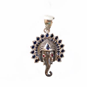 May include: Silver pendant featuring a detailed depiction of the Hindu deity Ganesha, with blue and red gemstones. The pendant is surrounded by a halo of smaller blue gemstones.
