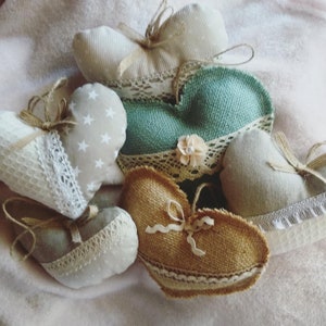 Può includere: Una collezione di sei cuscini a forma di cuore fatti a mano in diversi tessuti, tra cui juta, lino e pizzo. I cuscini sono decorati con fiocchi e bordi in pizzo.