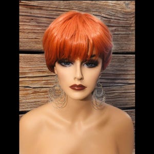 May include: A short, bright orange wig with blunt bangs. The wig is styled in a pixie cut and is worn on a mannequin head.
