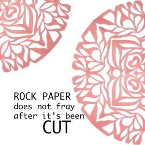 May include: Two pink paper snowflakes with intricate cut-out designs. The text "ROCK PAPER does not fray after it's been CUT" is written below the snowflakes.