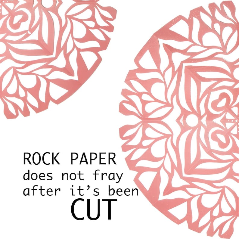 May include: Two pink paper snowflakes with intricate cut-out designs. The text "ROCK PAPER does not fray after it's been CUT" is written below the snowflakes.