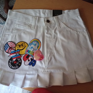 May include: White denim skirt with a ruffled hem and various colorful patches, including a smiley face, a unicorn, a star, and a heart.
