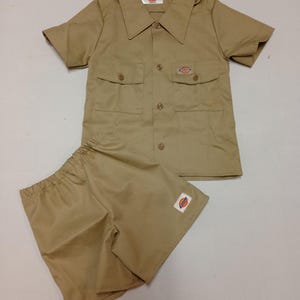 May include: A tan short-sleeved shirt and matching shorts set. The shirt has two chest pockets and a collar. Both pieces have a small logo patch. The set is made of a durable, workwear-style fabric.