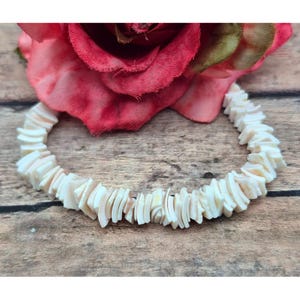 May include: A natural-toned shell bracelet with a segmented design. The bracelet is made of small, irregular shell pieces. A large, red rose is in the background, resting on a wooden surface.