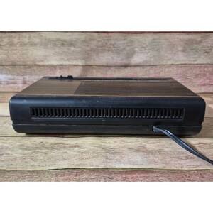 K&ouml;nnte beinhalten: Ein schwarzes und braunes Vintage-Elektronikger&auml;t mit Netzkabel. Das Ger&auml;t hat eine rechteckige Form mit horizontalen Linien auf der Oberseite und einem L&uuml;ftungsschlitz auf der Vorderseite. Es steht auf einer Holzoberfl&auml;che.