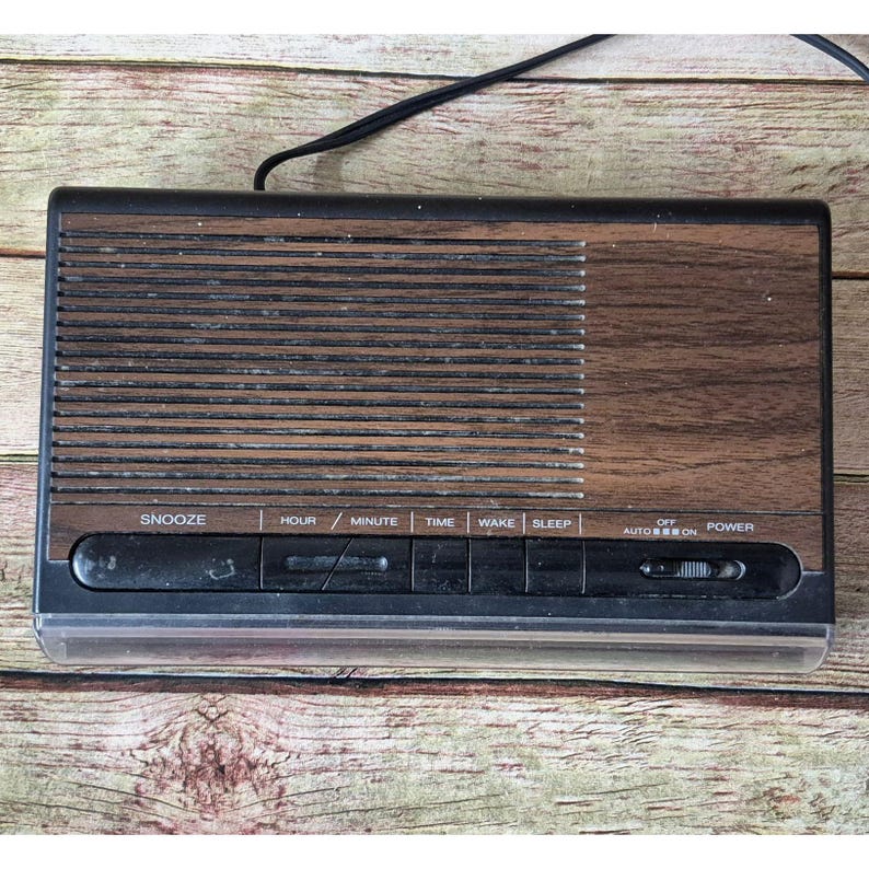 K&ouml;nnte beinhalten: Ein Vintage-Digitalwecker mit Holzmaserung auf der Frontplatte. Die Uhr hat einen schwarzen Kunststoffrahmen und eine Reihe von Tasten mit der Aufschrift "SNOOZE", "HOUR", "MINUTE", "TIME", "WAKE" und "SLEEP". Der Netzschalter ist mit "OFF AUTO ON" beschriftet.