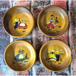 May include: Set of four wooden bowls with hand-painted Dutch scenes. Each bowl features a different scene with figures in traditional clothing, windmills, and other Dutch elements. The bowls are made of light-colored wood and have a glossy finish.