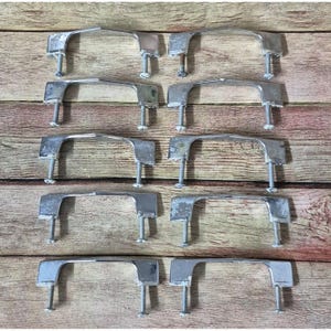 May include: A collection of ten vintage metal cabinet handles. Each handle is silver-toned, with a curved bar and two screw posts for mounting. The handles are arranged on a wooden surface, showcasing their design and potential for restoration projects.