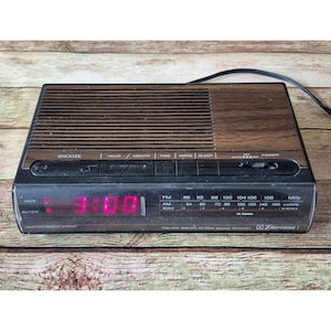 K&ouml;nnte beinhalten: Ein Vintage Emerson FM/AM Digital-Radiowecker mit Holzmaserung und schwarzem Sockel. Die Digitalanzeige zeigt die Uhrzeit 3:00 in leuchtend rosa Ziffern an. Funktionen: Schlummerfunktion, Zeit, Wecken und Schlaf.