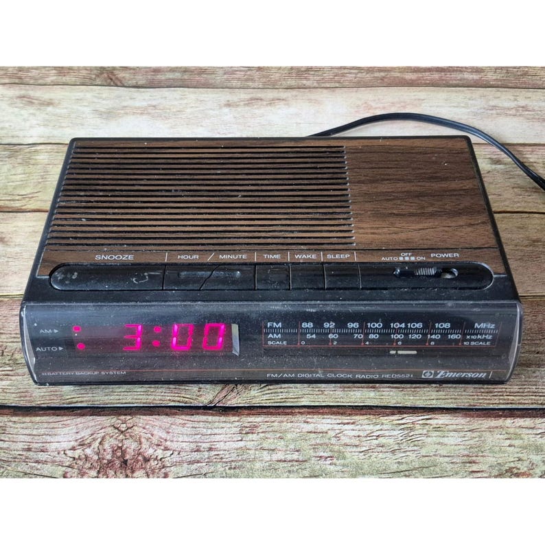 K&ouml;nnte beinhalten: Ein Vintage Emerson FM/AM Digital-Radiowecker mit Holzmaserung und schwarzem Sockel. Die Digitalanzeige zeigt die Uhrzeit 3:00 in leuchtend rosa Ziffern an. Funktionen: Schlummerfunktion, Zeit, Wecken und Schlaf.