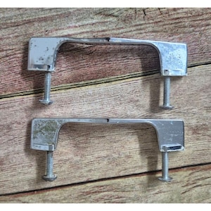 May include: Two chrome-colored cabinet handles with attached screws. The handles have a curved design and are slightly tarnished. The screws are silver and have a flat head. The handles are likely for a drawer or cabinet.
