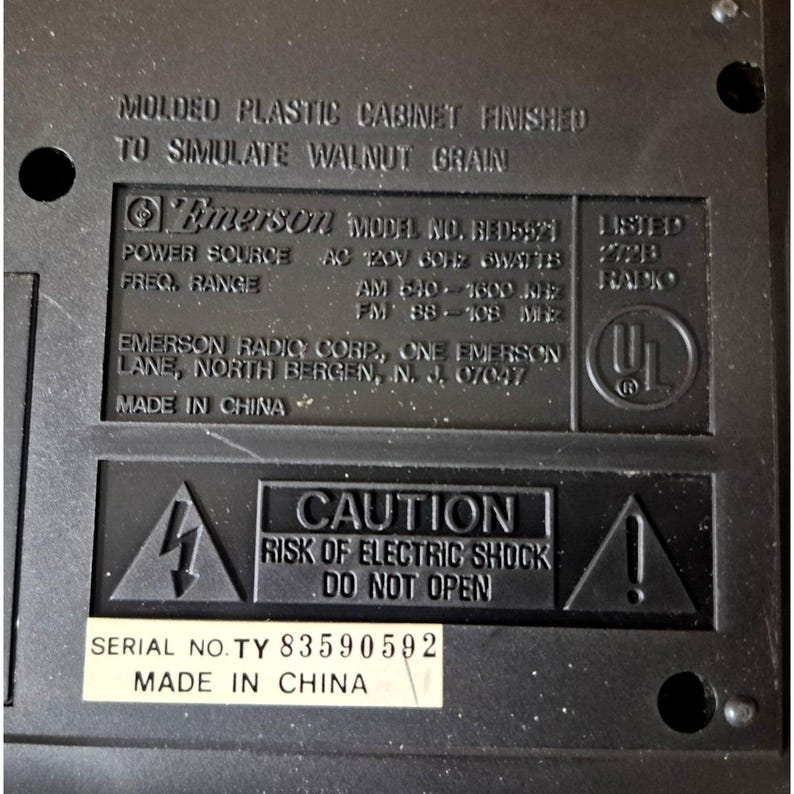 K&ouml;nnte beinhalten: Nahaufnahme der R&uuml;ckseite eines schwarzen Emerson-Radios. Auf dem Etikett steht "Geformtes Kunststoffgeh&auml;use, das Walnussholzmaserung simuliert". Es enth&auml;lt die Modellnummer, die Stromquelle, den Frequenzbereich und eine Warnung vor Stromschlag. Seriennummer ist TY 83590592.