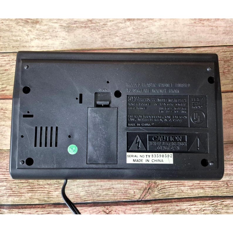 K&ouml;nnte beinhalten: Schwarzes Emerson-Radio mit einem Geh&auml;use aus geformtem Kunststoff, das eine Walnussmaserung simulieren soll. Die R&uuml;ckseite des Radios hat ein Batteriefach, eine Seriennummer und eine Warnung vor Stromschlag.