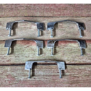 May include: Set of six vintage chrome cabinet pulls with mounting screws. The pulls have a curved handle design and are mounted on a wooden surface. The chrome finish shows signs of wear, adding to their vintage appeal.