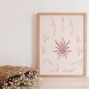 May include: A framed print with a pink background featuring a sun with a face and a circle of yoga poses around it. The poses are outlined in black and the sun is a darker shade of pink.