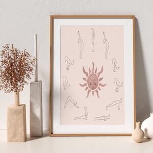 May include: A framed print with a pink background featuring a stylized sun with a face and a circle of yoga poses around it. The poses are outlined in black and the sun is a watercolor-like pink.