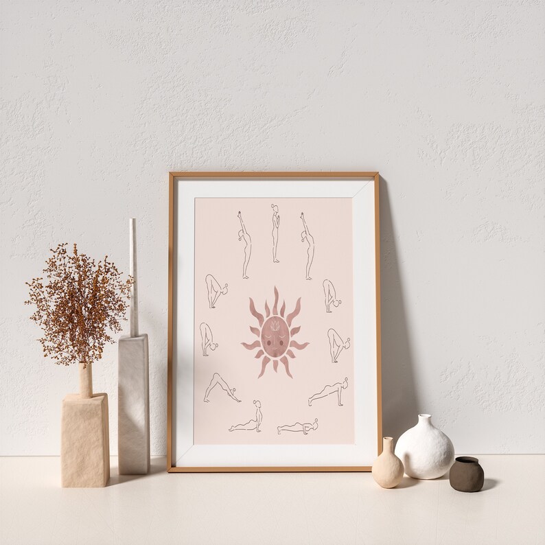 May include: A framed print with a pink background featuring a stylized sun with a face and a circle of yoga poses around it. The poses are outlined in black and the sun is a watercolor-like pink.