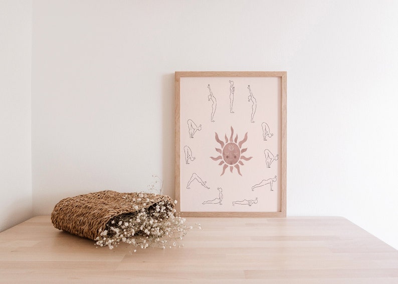 May include: A framed print with a pink background featuring a sun with a face and a circle of yoga poses around it. The poses are outlined in black and the sun is a darker shade of pink.