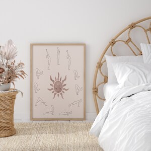May include: A framed print of a yoga sun salutation sequence with a sun illustration in the center. The print is in a light brown frame and is hanging on a white wall. The print is in a minimalist style with a light pink background and brown lines.