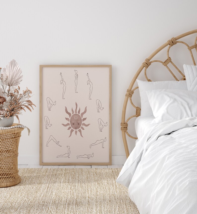 May include: A framed print of a yoga sun salutation sequence with a sun illustration in the center. The print is in a light brown frame and is hanging on a white wall. The print is in a minimalist style with a light pink background and brown lines.