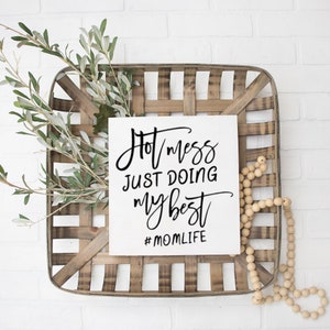 以下が含まれることがあります： 「Hot mess just doing my best #momlife」と黒の書道で書かれた白い看板。看板は木製の箱の中に置かれ、木製のビーズの紐が置かれている。