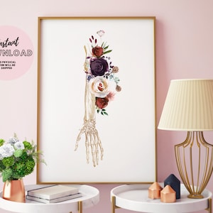 May include: A printable art print featuring a gold skeletal hand with a bouquet of burgundy, white, and purple flowers. The print is framed and displayed on a table with a lamp and other decor.