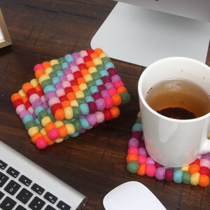 May include: A set of four colorful felt coasters with a rainbow pattern. The coasters are made of small, round felt balls that are woven together. A white mug with tea is sitting on one of the coasters.