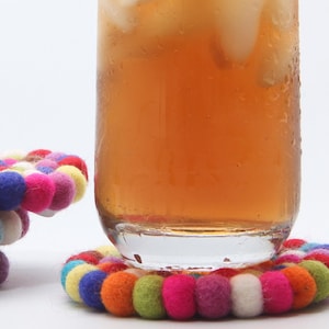 May include: A glass of iced tea sits on a colorful felt ball coaster. The coaster is made of small, round felt balls in various colors, including pink, blue, yellow, and orange. Another coaster is stacked nearby.