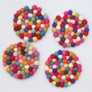 May include: Four round coasters made of colorful felt balls. The coasters are arranged in a square, each featuring a unique mix of red, orange, yellow, green, blue, purple, and white balls. The background is plain white.