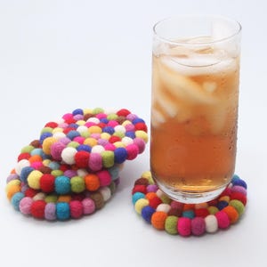 May include: A glass of iced tea sits on a colorful, round felt coaster. Several other coasters are stacked nearby. The coasters are made of small, brightly colored felt balls in shades of red, pink, yellow, blue, and green.