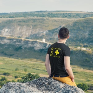 May include: A person wearing a black t-shirt with a yellow graphic that says "SAR Search Rescue" and a yellow plus sign. The person is sitting on a rock overlooking a valley.