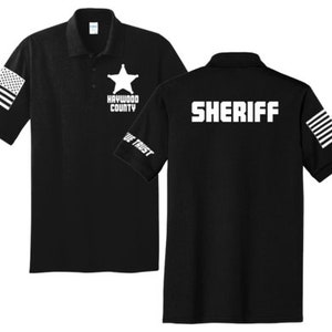 May include: Black polo shirt with a white star and the text "Haywood County" on the left chest and the word "SHERIFF" on the back. The shirt has a white American flag design on both sleeves.