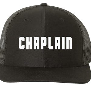 May include: Black and gray trucker hat with a white mesh back. The front of the hat has the word "CHAPLAIN" in white block letters.