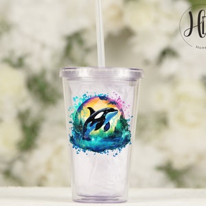 May include: A clear plastic tumbler with a colorful design featuring an orca whale jumping out of the water. The design includes a watercolor effect with shades of blue, green, and pink. The tumbler has a straw.