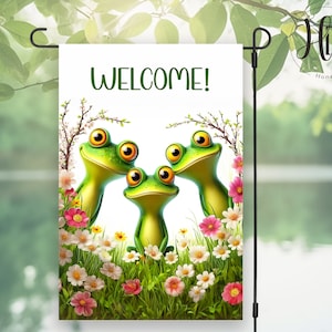 Puede incluir: Una bandera de jardín de bienvenida con tres ranas de dibujos animados en un campo de flores. Las ranas son verdes con ojos grandes y miran hacia arriba. La bandera es blanca con la palabra "WELCOME!" en letras verdes.