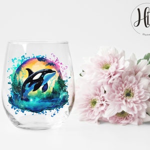 May include: A clear glass wine glass with a colorful watercolor design of an orca whale jumping out of the water. The design includes a sunset, trees, and water splashes.