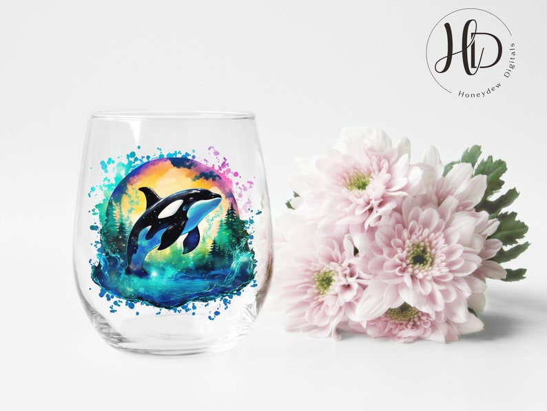 May include: A clear glass wine glass with a colorful watercolor design of an orca whale jumping out of the water. The design includes a sunset, trees, and water splashes.