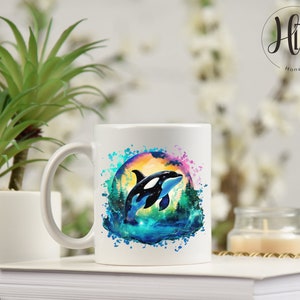 May include: White ceramic mug with a colorful watercolor design of an orca jumping out of the water. The orca is black and white, and the water is blue and green. The background is a sunset with pink and orange colors.