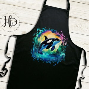 May include: Black kitchen apron with a colorful watercolor design of an orca whale jumping out of the water. The design has a rainbow color scheme with a dark blue background.