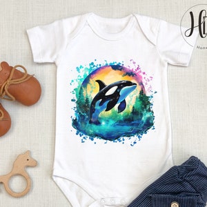 May include: White baby onesie with a watercolor design of an orca whale jumping out of the water. The design features a colorful background with splashes of blue, green, and purple.