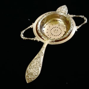 May include: Ornate silver tea strainer with a perforated bowl, two handles, and a decorative handle. The strainer sits on a round base with a raised rim. The handle and rim feature intricate floral patterns, adding to its vintage charm.